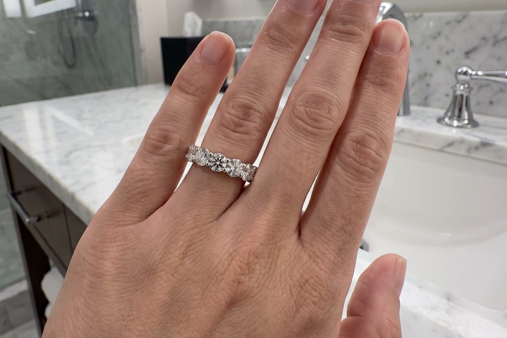 Round lab grown diamond eternity band in 14K white gold worn on a hand at a marble vanity, Draco Diamond