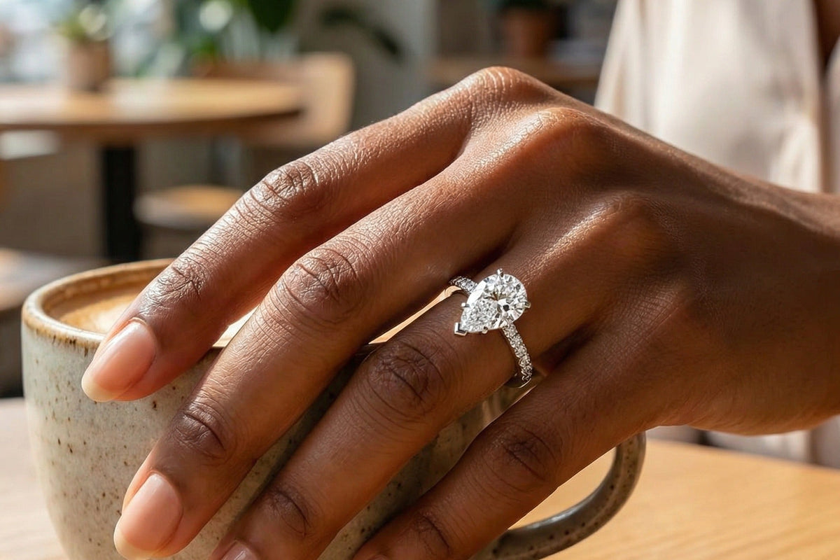 Hand holding a ceramic mug at a cafe table wearing a lab grown diamond engagement ring with a hidden halo under the centre stone, Draco Diamond